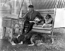 A man sits on a swing, writing on a crude desk. Two dogs, Rough Collies, sit on his left.  Another lies on the ground at his feet.