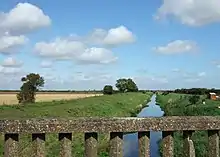 From the parapet of a lichen covered concrete bridge we are looking along the line of the navigation, which stretches dead straight to the horizon.  It is well filled with placid water. There are wide, grass-covered embankments on each side, with wide grassed tracks on top.  To the side are fields of ripened wheat, the nearest just harvested.  A few oak trees stud the distant embankments.  It is a glorious summer day with a deep blue Lincolnshire 'big sky', studded with small fluffy white clouds.